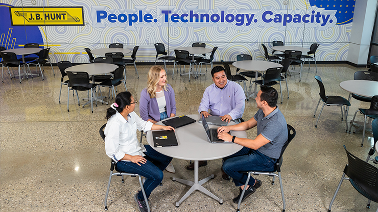  Shelly Simpson sitting with three people at a table in front of a wall that has the J.B. Hunt logo and says "People Technology. Capacity."