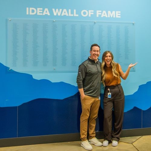 Two J.B. Hunt employees are smiling in front of a blue wall that reads 'Idea Wall of Fame' with hundreds of names listed below.