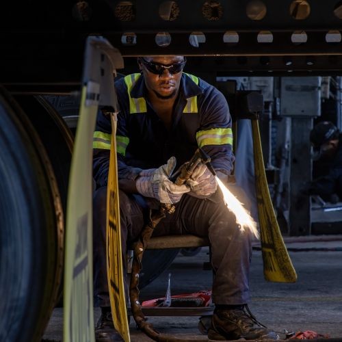 J.B. Hunt maintenance technician is sitting on a stool in a reflective jumpsuit and safety glasses. He is about to begin welding on the underneath of a semi-truck trailer.