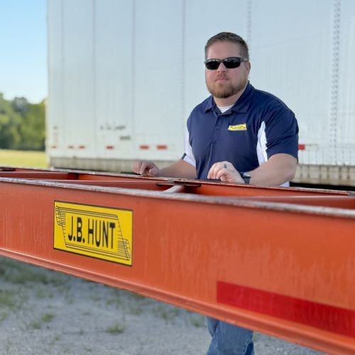 J.B. Hunt employee standing behind an orange J.B. Hunt Intermodal chassis, while wearing a navy blue polo and sunglasses.