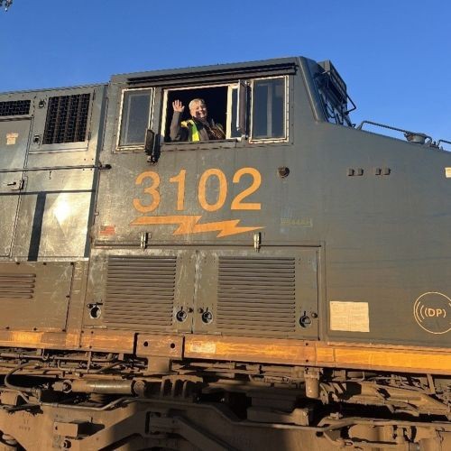 J.B. Hunt employee waving out of a train car window in a neon yellow safety vest. The train is black with the numbers 3102 in orange with an orange lightning bolt underneath."