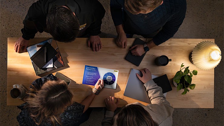 Top-down view of four team members collaborating at a wooden table, reviewing a document and using digital devices, with a warm desk lamp and small plant nearby.