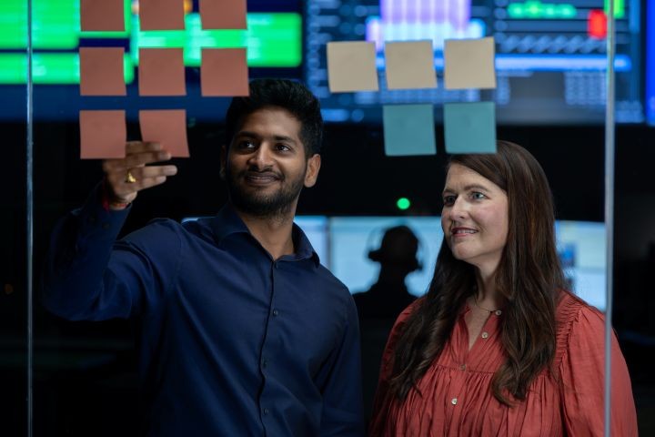 Two people working together, placing sticky notes on a glass wall in front of digital screens displaying data.