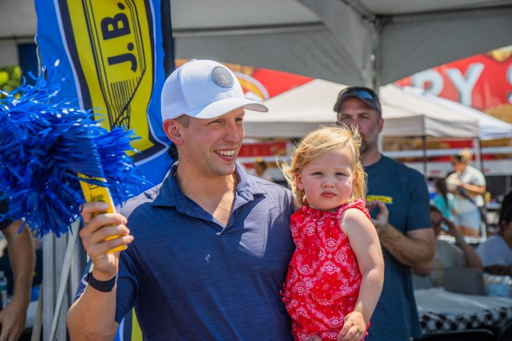 J.B. Hunt employee in a white hat and blue shirt holds a child and pom-pom on a sunny day.