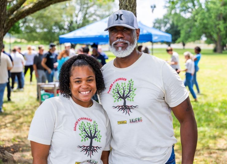 A man and a woman stand smiling in a grassy area with an event happening behind them.