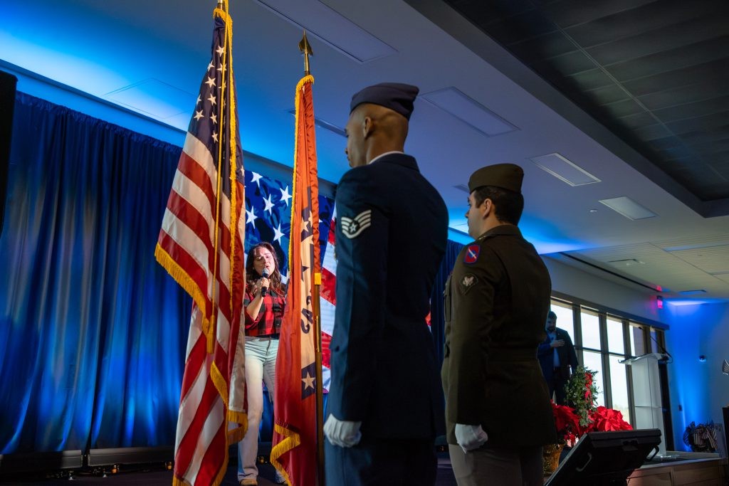 Two military members standing in front of the U.S. and Arkansas flag as someone sings on stage.