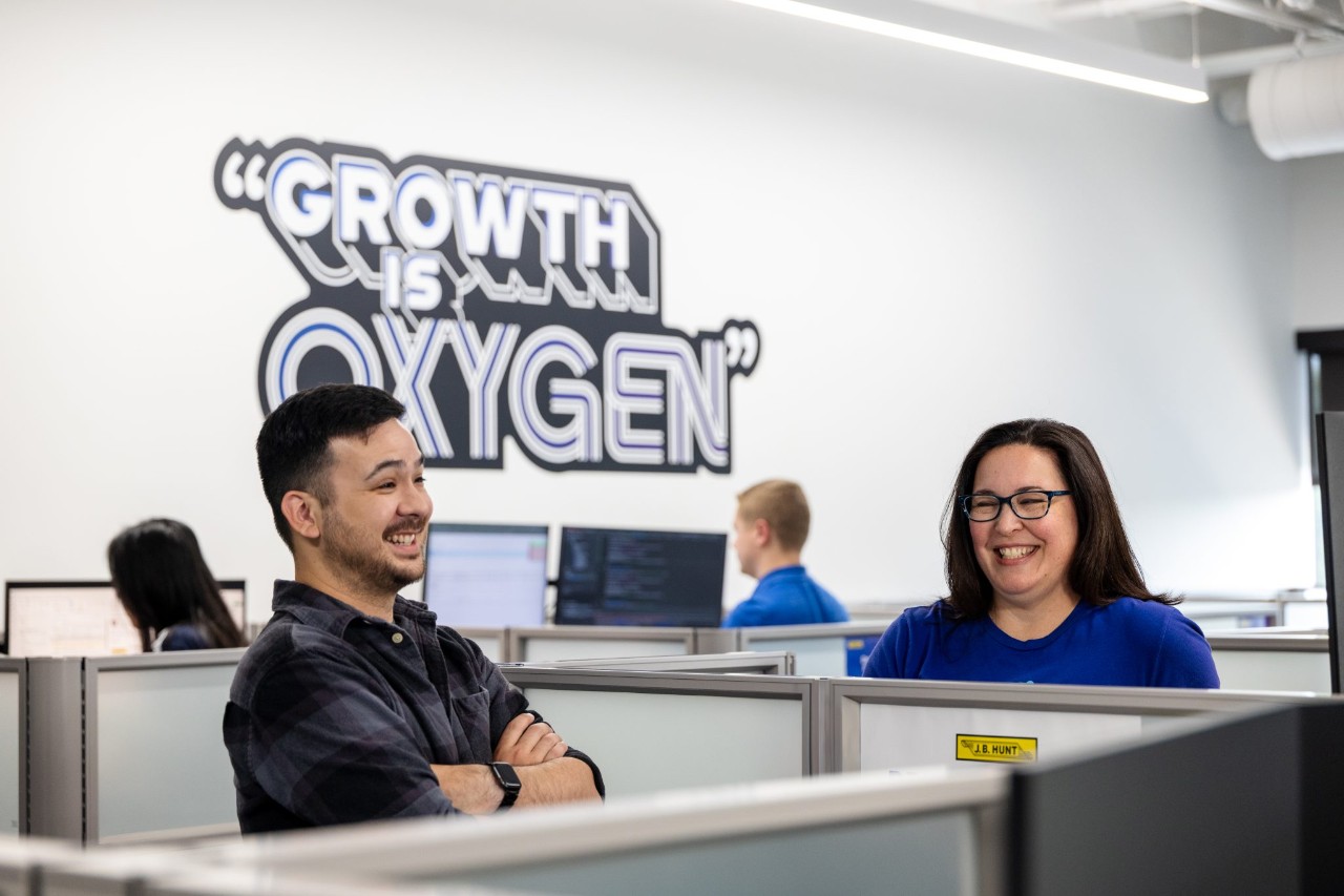 Employees conversing with each other at their cubicles in front of a wall decal that reads, "Growth is Oxygen".
