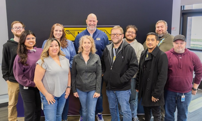 A group of eleven J.B. Hunt employees smiling in front of the yellow J.B. Hunt scroll logo.