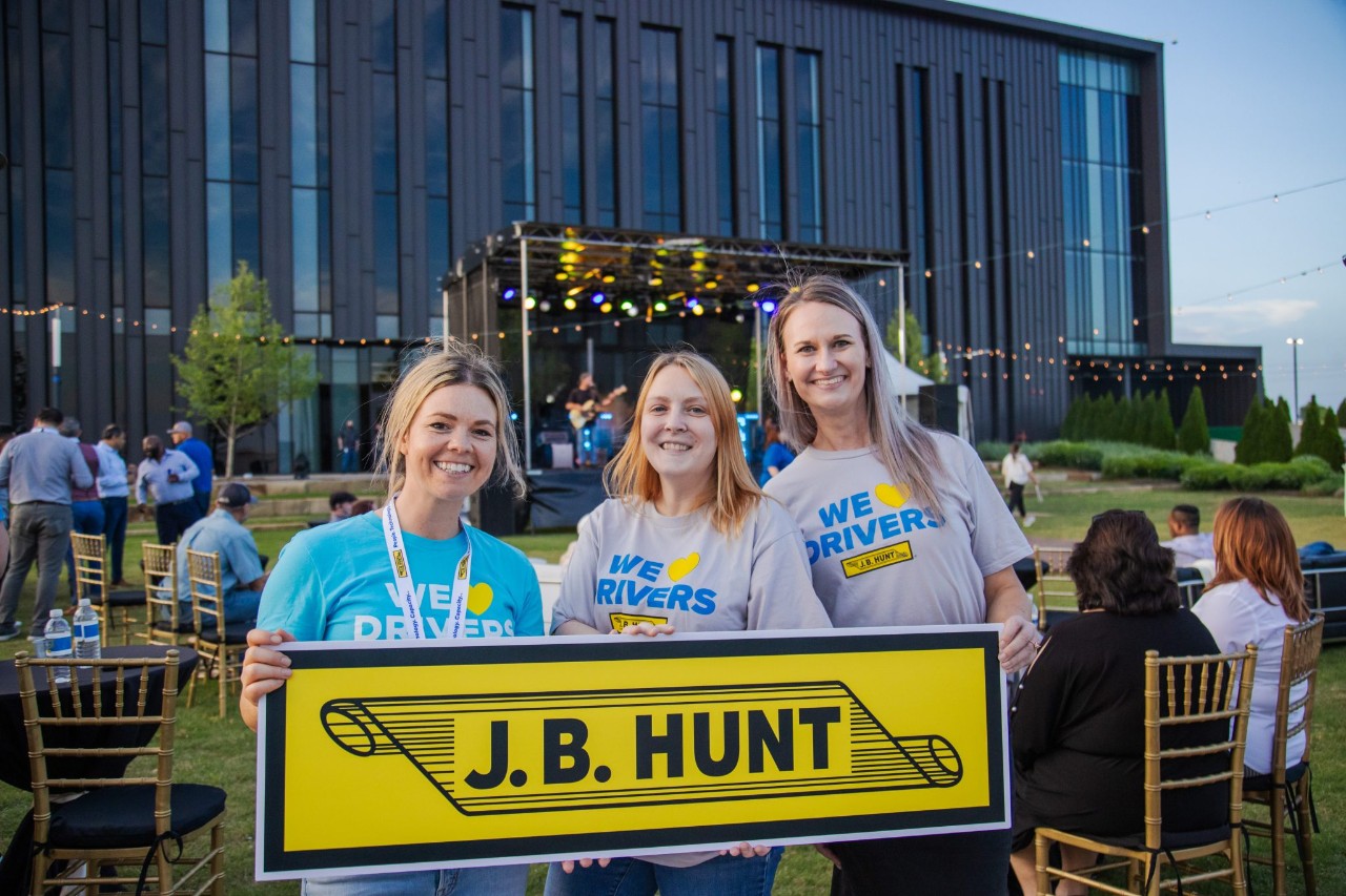 three women smiling wearing shirts that say we love drivers and holding the J.B. Hunt scroll logo