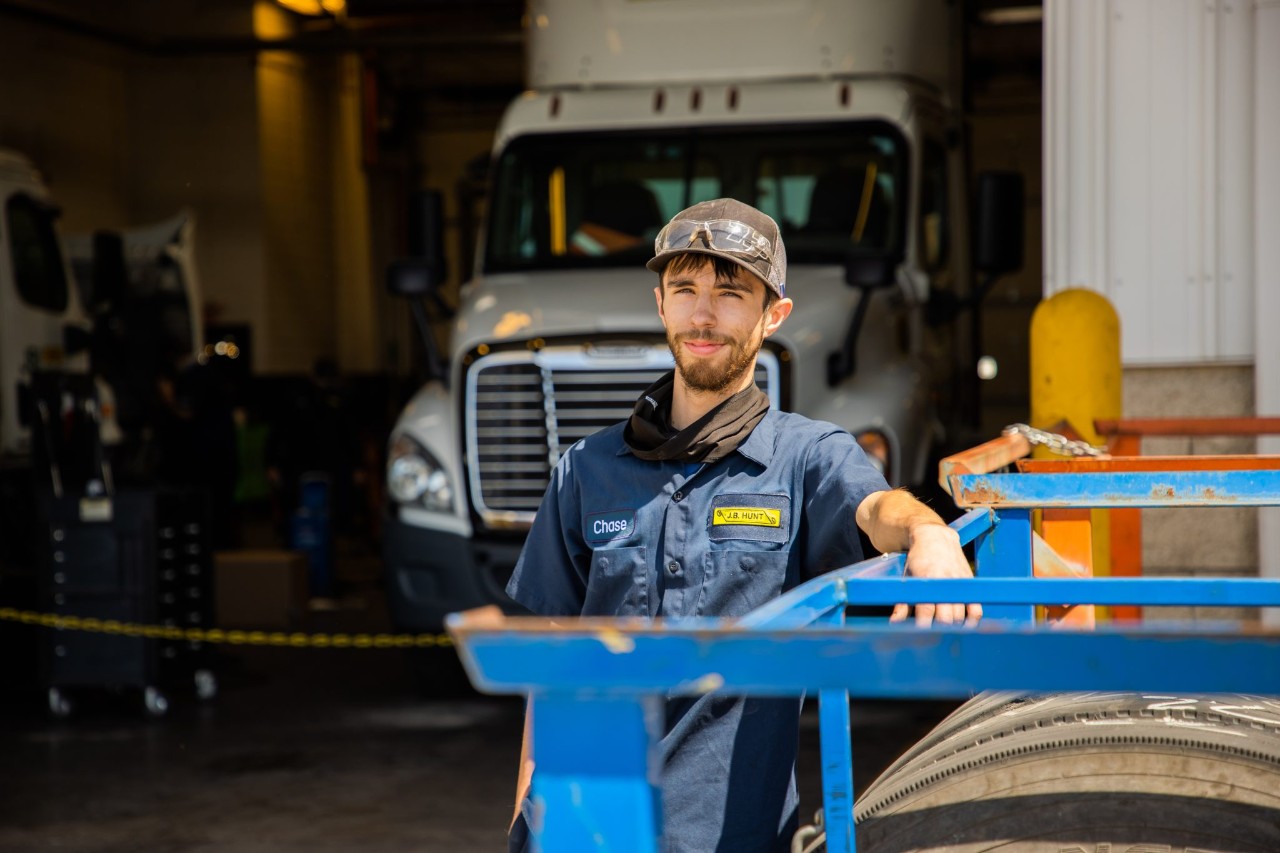 Male, maintenance technician leans on tire with semi-truck in the background.