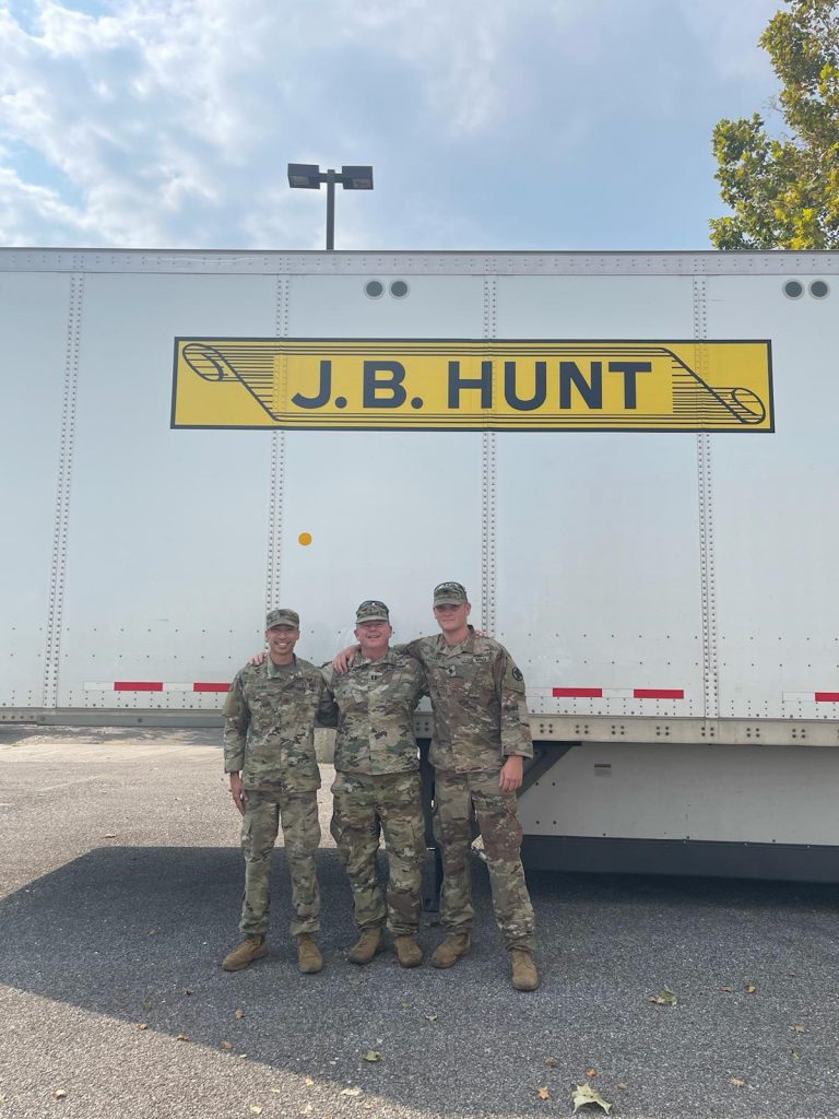 Nicholas (right) and two other J.B. Hunt military employees with arms on shoulders, in uniform, in front of J.B. Hunt truck.