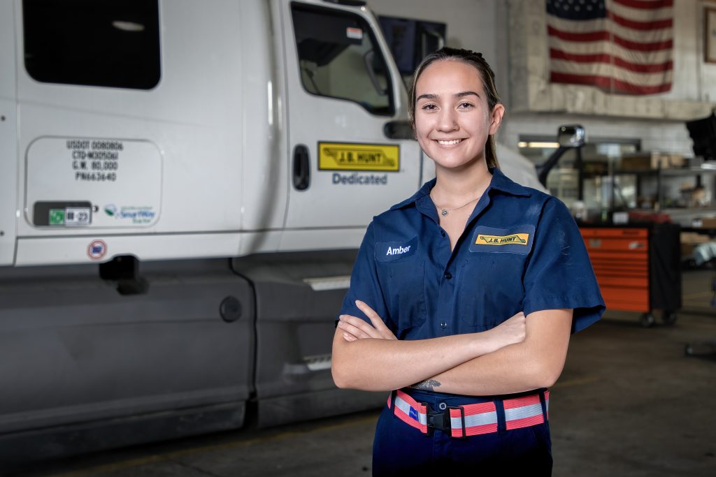 Woman maintenance technician wearing J.B. Hunt uniform stands with arms crossed in front of semi-truck and American flag.