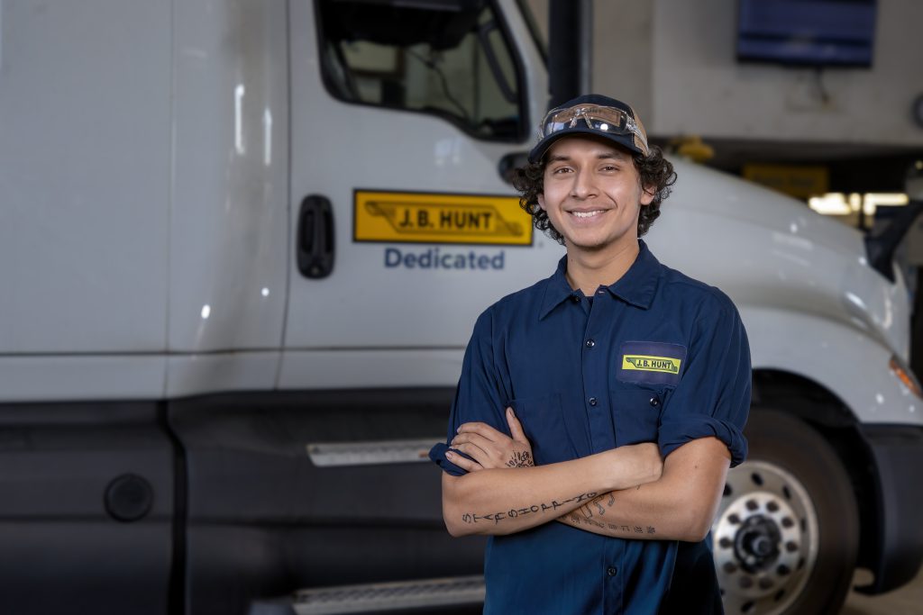 Young, male maintenance technician stands smiling in front of J.B. Hunt semi-truck. 