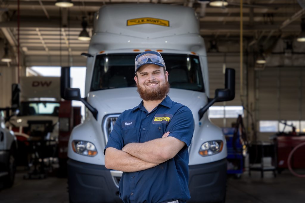 Male, maintenance technician with a beard and glasses on his head stands in front of forward-facing semi-truck with his arms crossed as he smiles. 