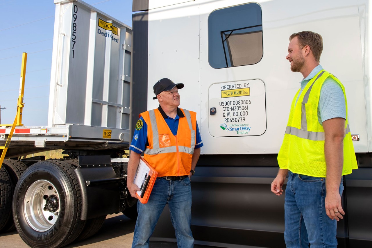 A J.B. Hunt employee in a neon yellow vest talks with a truck driver wearing an orange safety vest next to a semi-truck.