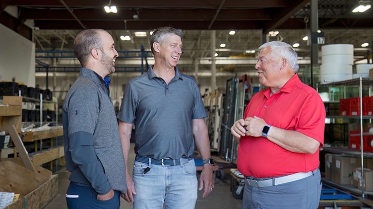 Two J.B. Hunt employees in casual work attire having a friendly conversation with a customer wearing a red polo shirt inside a warehouse. The setting includes industrial shelving and equipment.