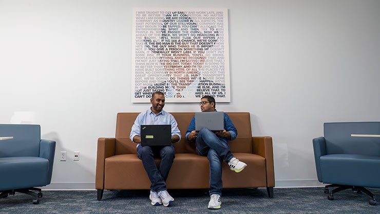 Two professionals sitting on a brown couch in a modern office, working on laptops and engaging in discussion. A large artistic print with text is displayed on the wall behind them.