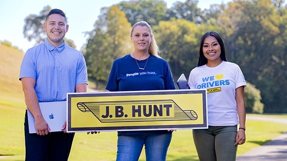 Three j.b. hunt employees standing outdoors holding a J.B. Hunt sign, showcasing team pride, with one person holding a laptop and wearing shirts that display supportive slogans.
