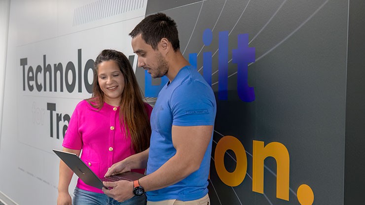 A male and female employee standing in a hallway, looking at a laptop together. 