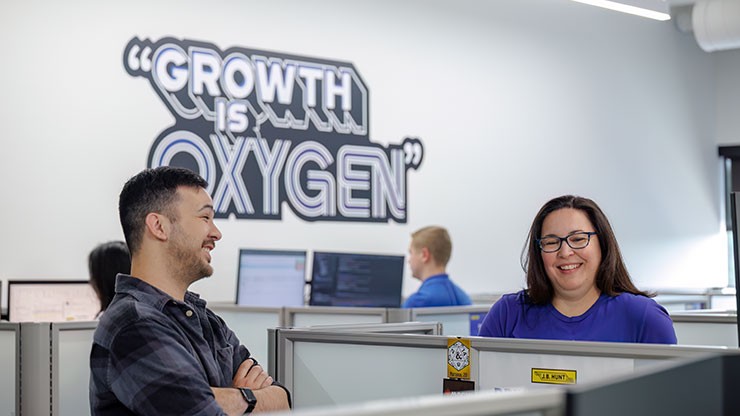 Employees in an office space smiling and conversing near cubicles, with a motivational quote ‘Growth is Oxygen’ displayed on the wall in the background.