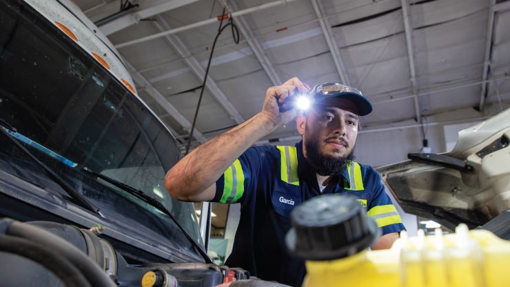 Mechanic inspecting a vehicle engine with a flashlight in a garage, wearing a uniform with reflective safety stripes.