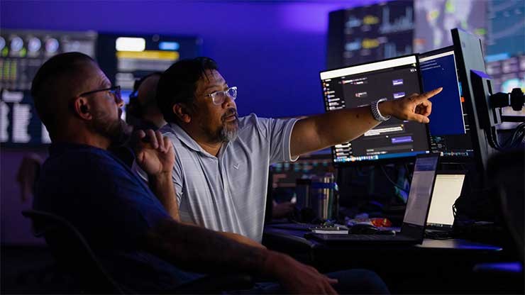 Two men in front of many screens. One points to something on a screen